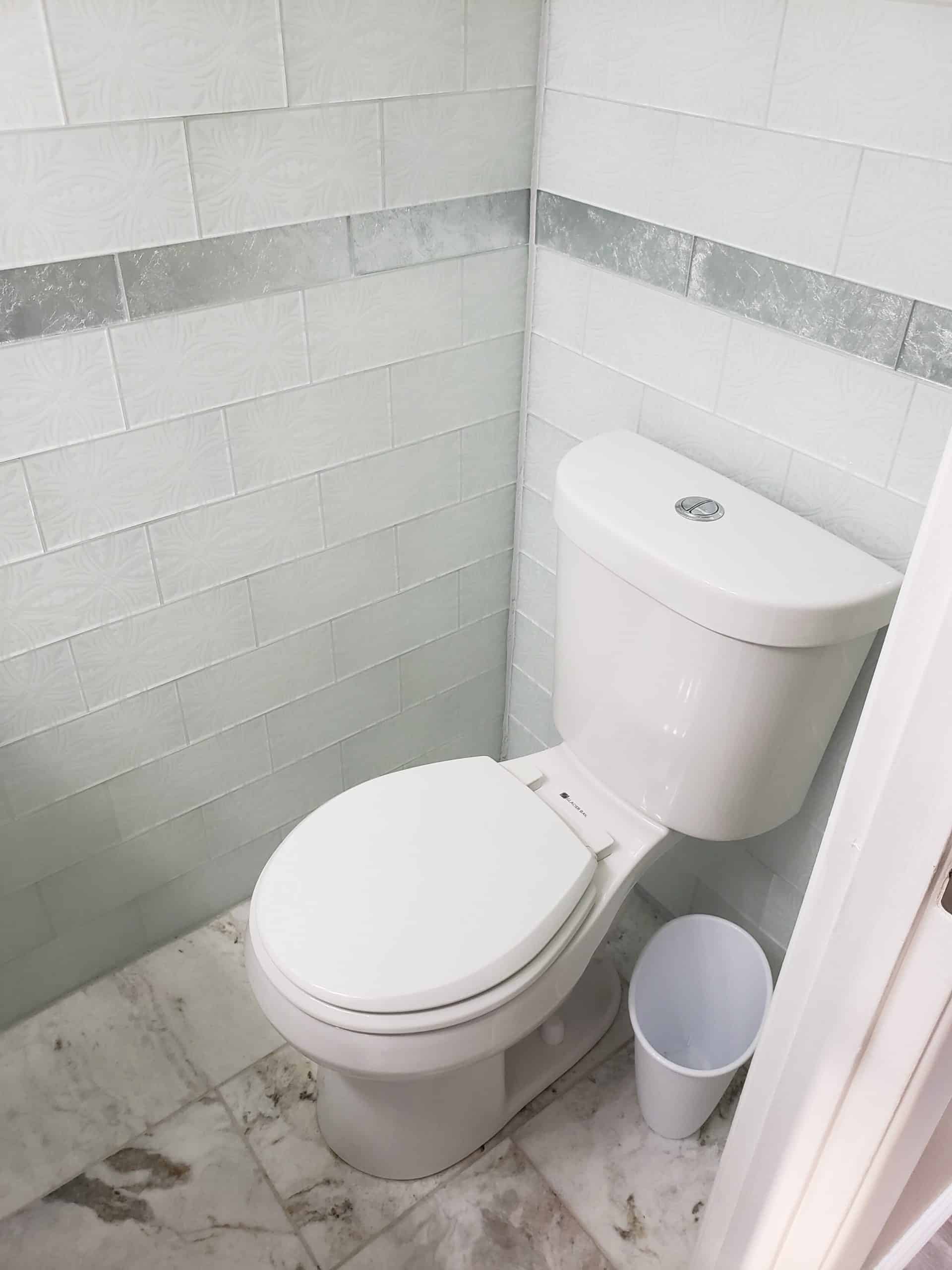 Half bathroom toilet in front of light gray tile accent wall